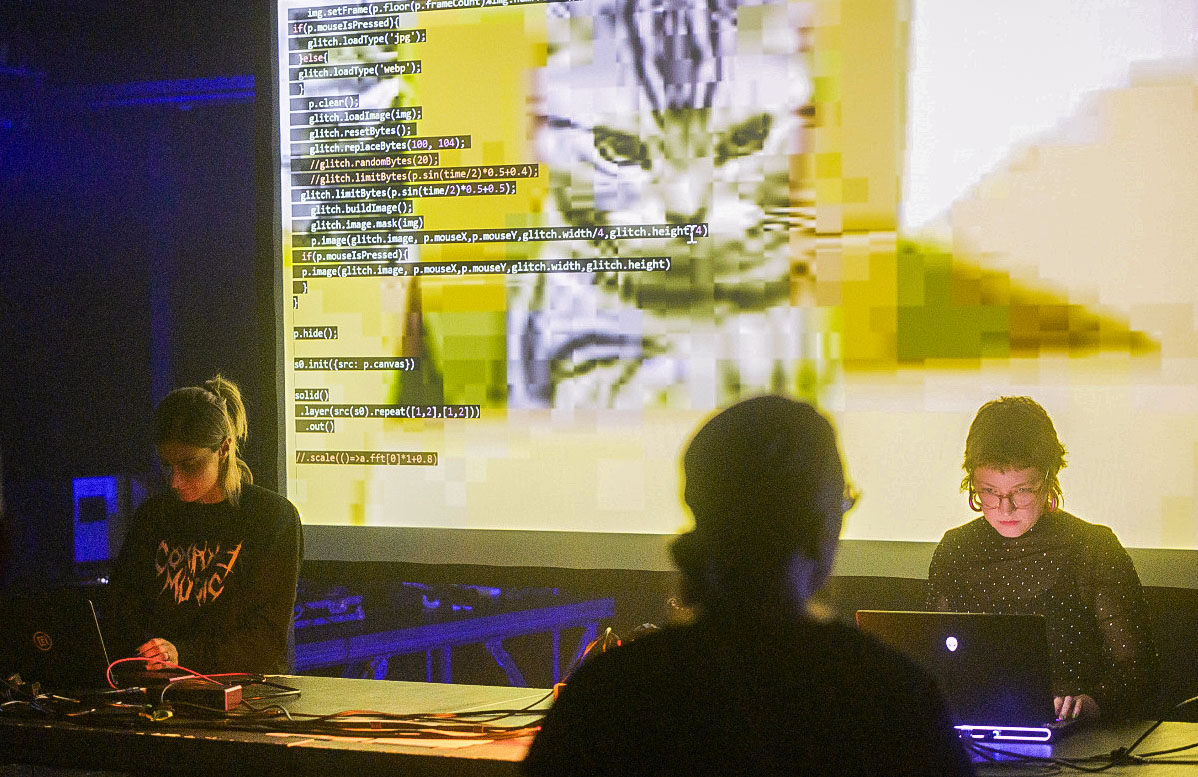 two performers at a table with a disapproving cat covered in code on a screen behind them
